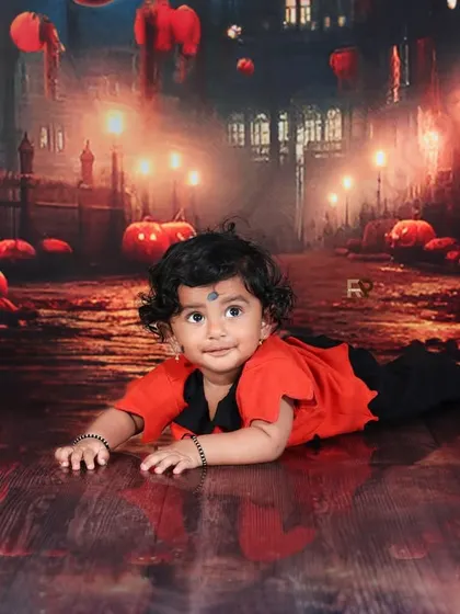 A spooky and sweet Halloween-themed photo. A baby in a cute devil costume crawls on a floor with pumpkin reflections, set against a backdrop of a haunted street.
