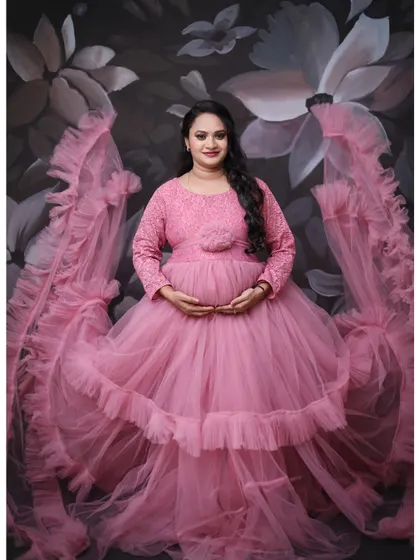 A full-length portrait showing the dramatic flair of a pink ruffled gown with extended fabric wings, set against a floral painted backdrop.