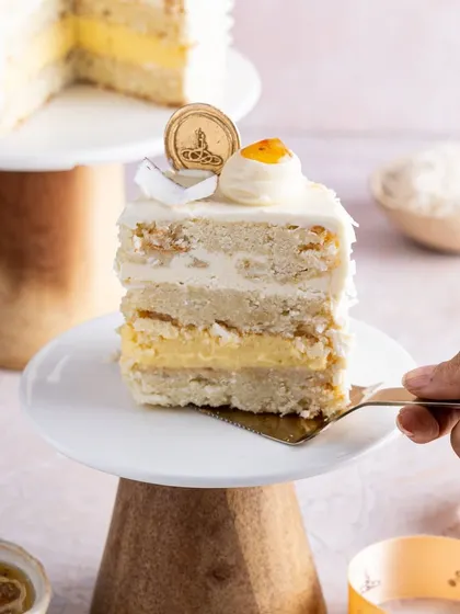 A slice of our tropical Coconut and Passionfruit cake on a dessert stand. This cake features layers of fluffy coconut sponge, a bright passion fruit cremeux, and a smooth coconut buttercream, perfect for a taste of summer.