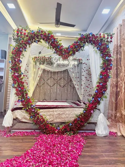 A stunning wedding night setup featuring a large heart-shaped arch of red flowers framing the bed. A path of rose petals leads to the bed.