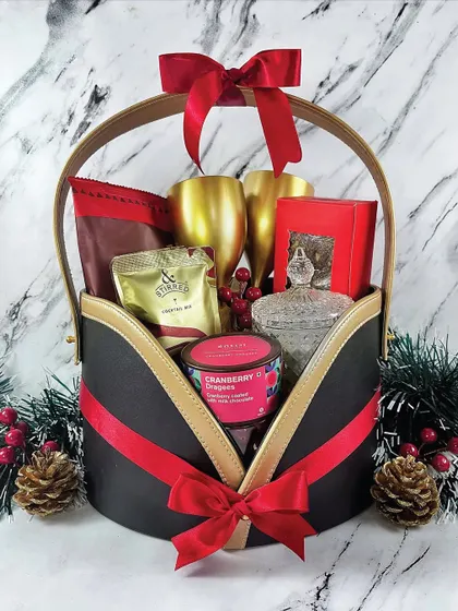 A stunning holiday gift basket in a unique black, red, and gold container. It's filled with two golden goblets, a cocktail mix, and other gourmet goodies for a festive celebration.