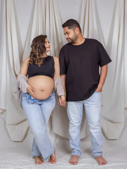 A full-length shot of a casual maternity pose. The couple holds hands, dressed in comfortable denim and black, standing against a simple white draped background for a clean and modern feel.