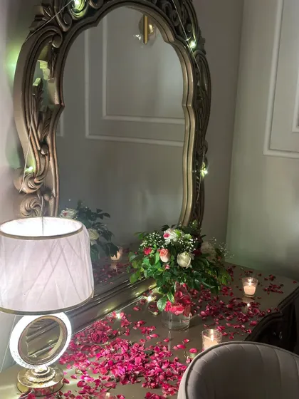 A detail shot of the romantic room decor. A dressing table is adorned with a flower bouquet, scattered rose petals, and candles, creating a soft and intimate ambiance.