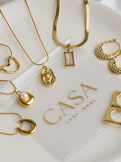 A collection of gold necklaces and earrings displayed on a branded plate against a soft white fabric. This group shot is great for showcasing a new collection or best-selling items.
