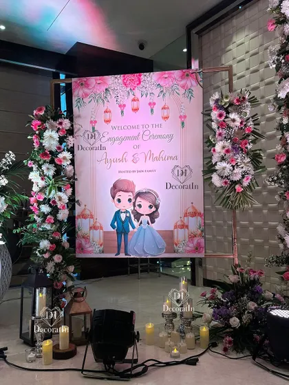A personalized welcome sign for Ayush and Mahima's engagement ceremony. The sign features a cute caricature of the couple and is framed by pillars of fresh pink and white flowers.