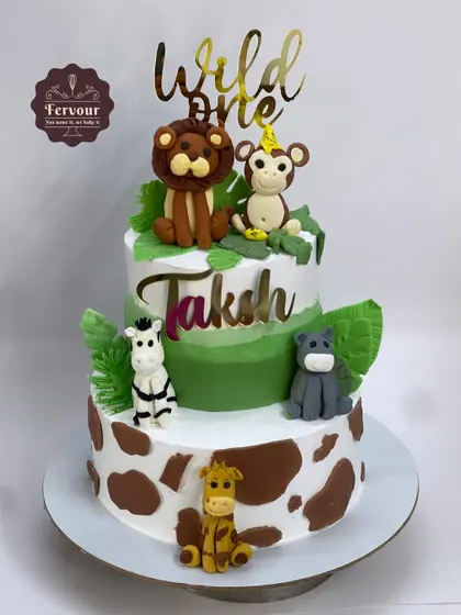 A "Wild One" animal-themed cake for a first birthday. This two-tier cake features adorable 3D fondant animals, a giraffe-print bottom tier, and a custom gold topper.