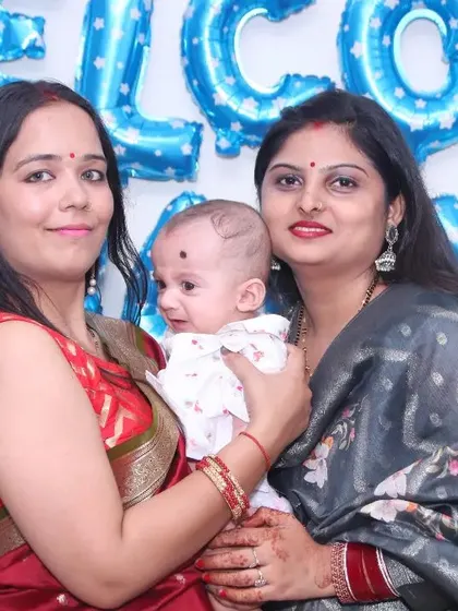 A photo of two women holding a newborn baby at a celebration with 'Welcome Baby Boy' balloons. We document these happy gatherings for new parents.