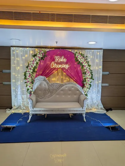 An elegant stage for a Roka ceremony at a banquet hall, designed with a floral ring, white and pink drapes, fairy lights, and a custom 'Roka Ceremony' neon sign.