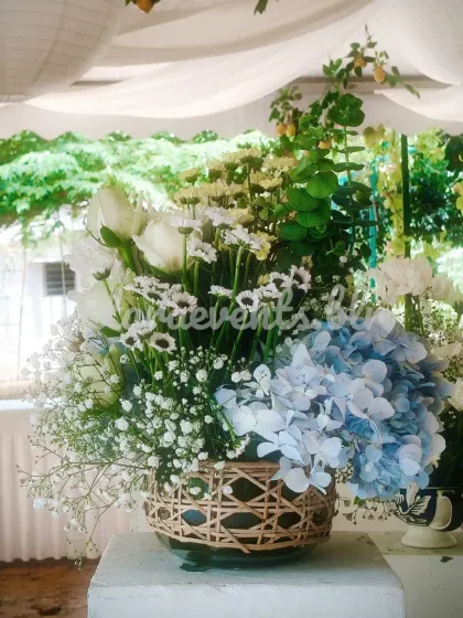 A beautiful floral arrangement from a Sicilian-themed event. The bouquet of white and blue flowers, including hydrangeas, is placed in a rustic woven vase, perfectly capturing the Mediterranean aesthetic.