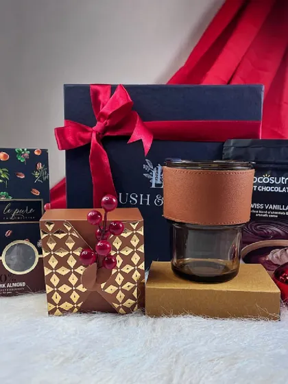Our 'Cozy at Home' hamper, presented with a deep red background. It includes dark almond chocolate, hot chocolate mix, and a glass tumbler, perfect for a relaxing evening.