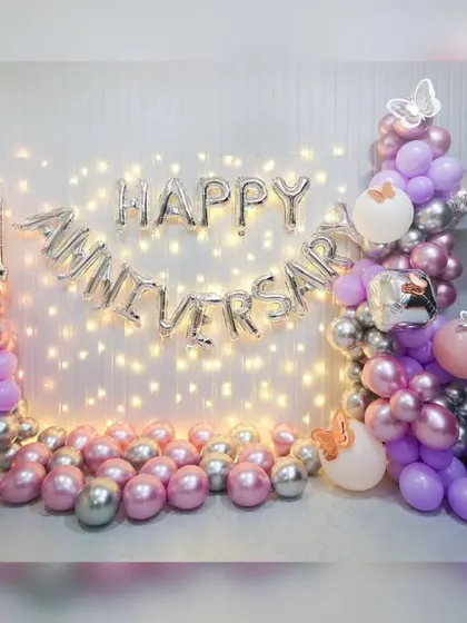 A first-anniversary decoration at home with a fairy light wall, 'Happy Anniversary' foil balloons, and a pink and silver balloon garland.