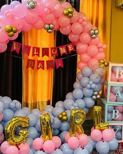 An "It's a Girl" welcome baby decoration. The setup includes a balloon arch in pink and blue, "GIRL" foil letters, and "BABY" blocks.