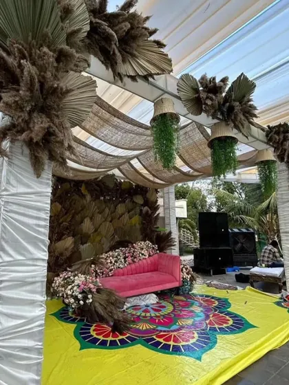 A wider view of the boho-chic Haldi stage, featuring a colorful mandala-printed floor, a pink lounge, and a backdrop of natural dried leaves under a tent.
