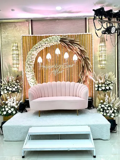 This is a seating area designed for a bohemian-themed Sangeet. The stage features a backdrop of gold fabric and dried palm leaves, with a "Happily Ever After" sign. The pink sofa and floral arrangements with pampas grass complete the chic, modern-boho look.