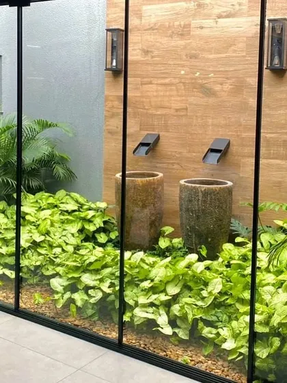 A modern indoor courtyard viewed through a glass wall. This design features a water element with two contemporary spouts, surrounded by a dense bed of syngonium plants, creating a serene and maintenance-friendly green space.