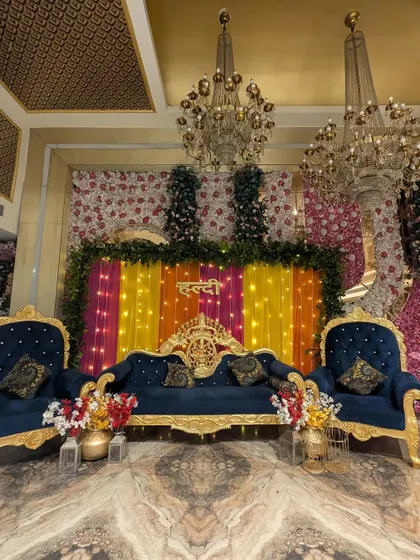 A royal Haldi stage decoration featuring a luxurious navy blue and gold sofa set. The backdrop is a vibrant mix of colorful drapes, a 'Haldi' sign, and a wall of flowers, creating a rich and opulent look.