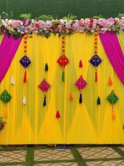 A bright and colorful backdrop perfect for a Haldi or Mehndi ceremony. This setup features a yellow curtain with pink and green drapes, decorated with traditional kite hangings.