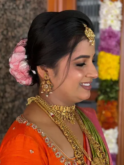 A smiling client showing her complete look, with a neat bun covered in a rose petal net that matches her saree.