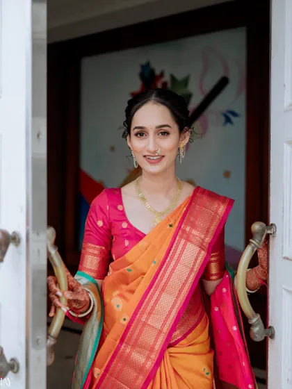 A welcoming and joyful portrait of the bride in a vibrant orange and pink saree. Her smile as she opens the doors creates a warm and inviting image, perfect for capturing the happiness of the occasion.