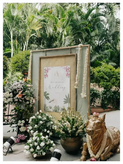 A beautiful welcome sign for a wedding, framed and adorned with fresh white flowers and a traditional brass Nandi statue. This creates an elegant and culturally rich entrance for guests.