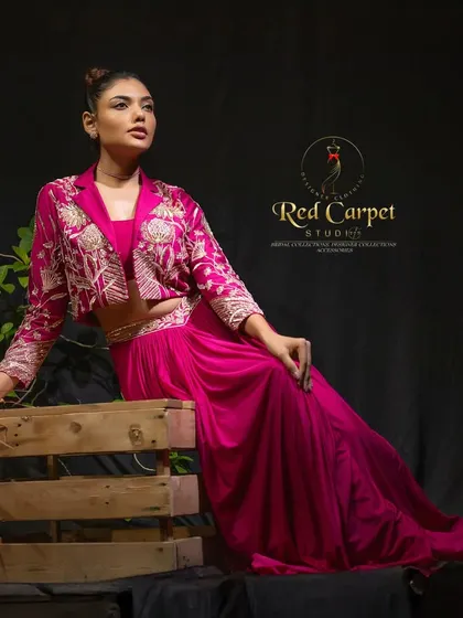 A model wearing a stunning fuchsia pink Indo-Western outfit. The ensemble includes a draped dhoti-style skirt and a bustier, paired with a cropped jacket featuring intricate gold floral embroidery.