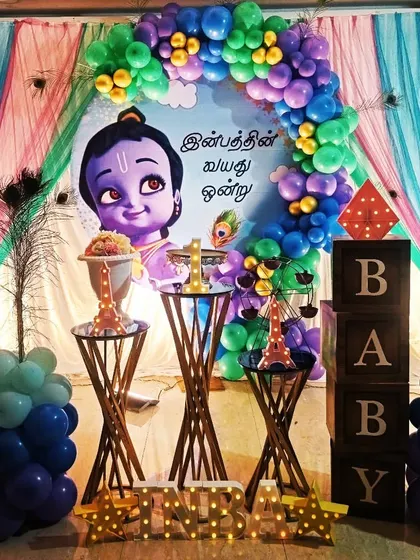 This is a custom Krishna-themed birthday party decoration. The setup features a playful illustration of little Krishna, peacock feather details, and a colorful balloon arch, making it a divine celebration for a child's special day.