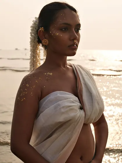 An ethereal beach shot where gold flakes on the model's skin catch the sunset light. The unique saree drape and the natural setting combine to create a powerful, artistic fashion statement.