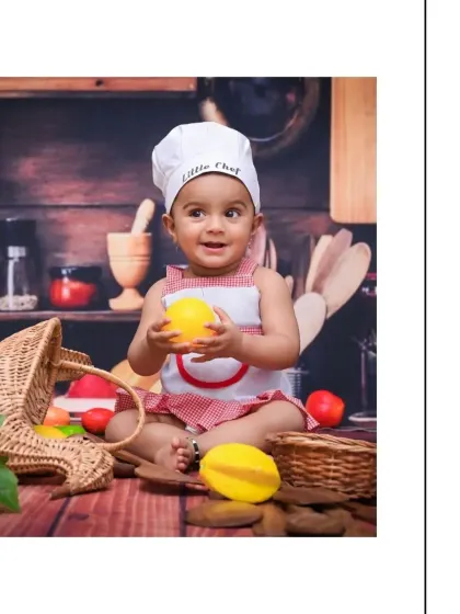 A happy baby in a chef's hat, holding toy lemons. The rustic wooden backdrop and colorful props create a warm and inviting atmosphere for this 'Little Chef' photoshoot.