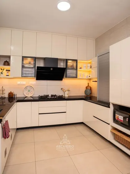 This modern kitchen combines glossy white cabinets with a black granite countertop and a sleek chimney, offering a high-contrast, contemporary feel.