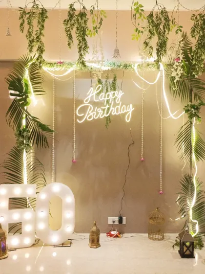 A beautiful bohemian-themed 60th birthday decoration. This elegant setup features a frame draped with greenery and fairy lights, a neon sign, and large marquee numbers.