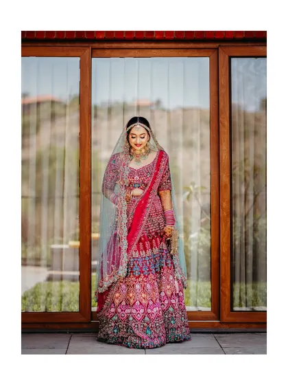 A classic full-length bridal portrait. The bride stands gracefully, showcasing her stunning pink lehenga and elegant poise against a modern backdrop.