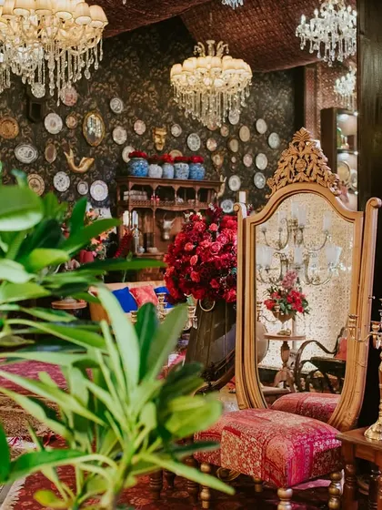A gilded antique mirror and vanity stool are framed by lush plants and crystal chandeliers, creating a quiet, elegant corner within the bustling, vibrant event space.