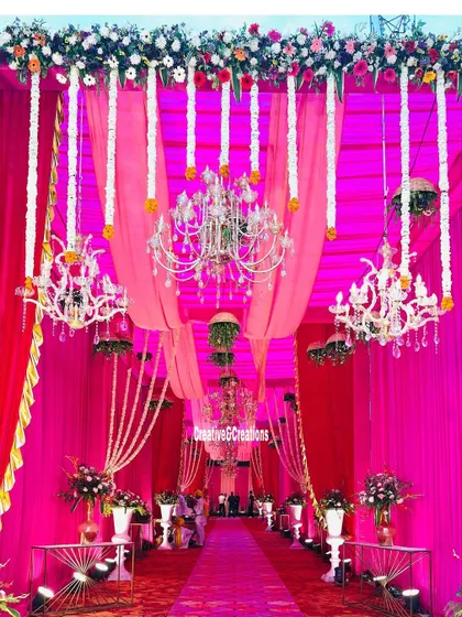 A stunning entrance saturated in vibrant pink. The ceiling is a mix of fabric draping, hanging floral strings, and crystal chandeliers, creating a truly grand and immersive experience.