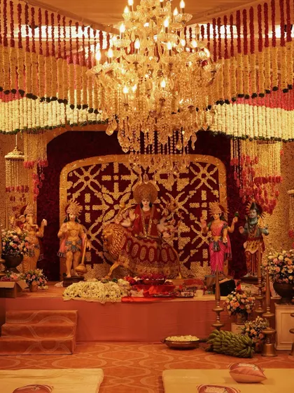 The sacred altar area for a Durga Puja, beautifully decorated with a backdrop of red and white florals and framed by the magnificent garland ceiling. The design is both grand and reverent.
