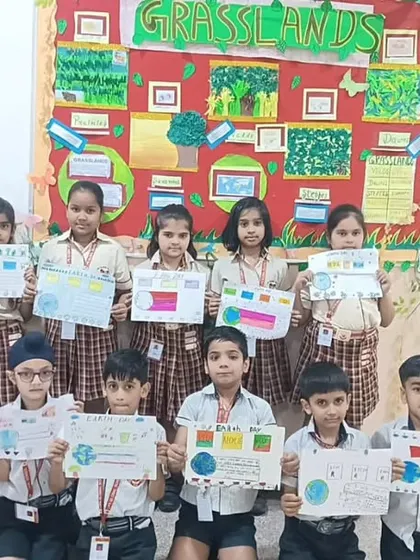 A group of students poses with their Earth Day posters in front of a bulletin board themed on grasslands, showcasing their classroom learning on ecosystems.