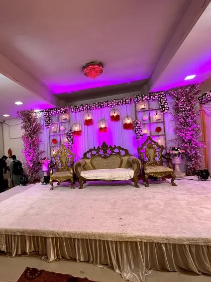 A grand wedding reception stage with ornate, royal style seating. The backdrop includes a grid of shelves with hanging birdcages and floral arrangements, lit with purple mood lighting.