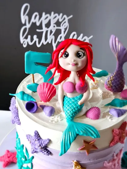 A close-up of the mermaid cake, showing the detailed, hand-sculpted mermaid figurine with her bright red hair. The surrounding edible shells and starfish complete the underwater scene.