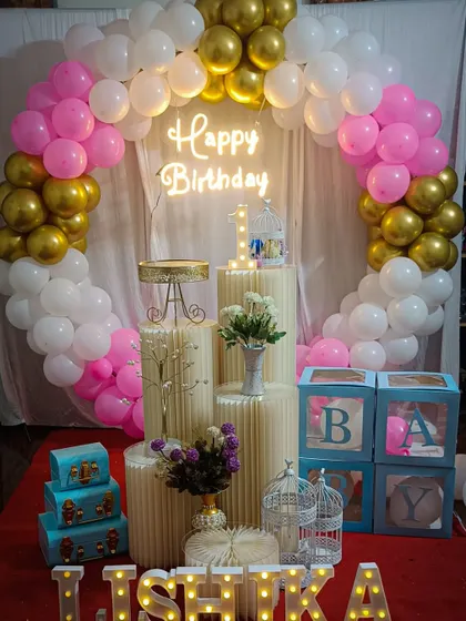 A sweet first birthday setup with a pink, white, and gold balloon arch, a 'Happy Birthday' neon sign, and charming details like birdcages and 'BABY' blocks.
