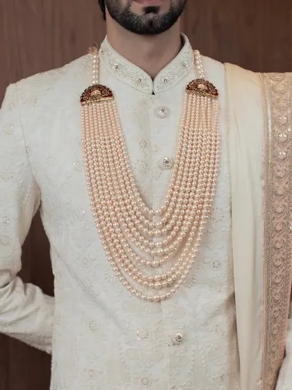 A close-up on the accessories that complete the royal look. The multi-layered pearl necklace with its ornate clasps perfectly complements the subtle texture of the ivory sherwani.