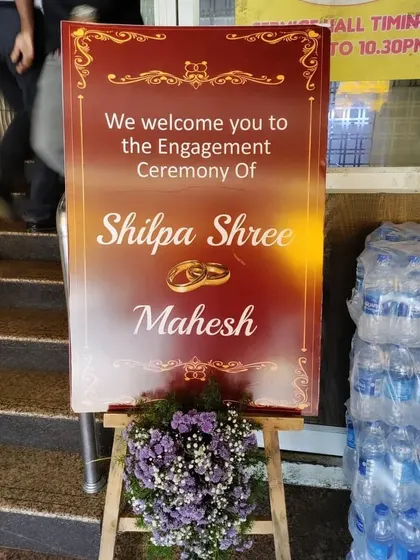 A classic welcome sign for an engagement ceremony, placed on an easel and decorated with a simple purple and white floral arrangement.