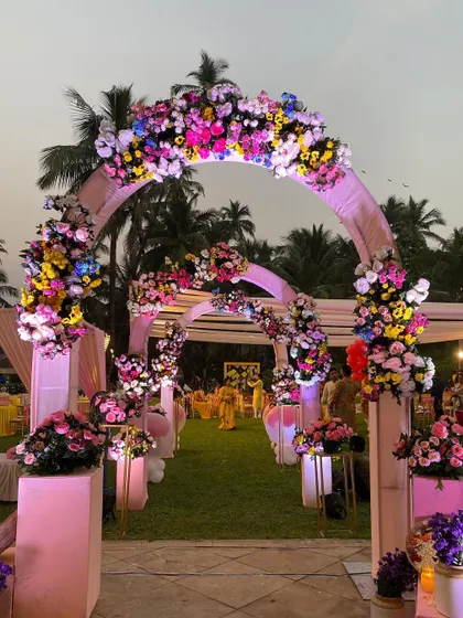 A vibrant, multi-colored floral archway for a daytime garden wedding. The mix of pinks, yellows, and blues creates a cheerful and festive entrance that is full of life.
