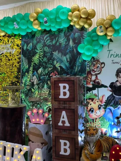 A detailed look at a jungle-themed birthday party. The decor includes a foliage backdrop, animal props like tigers and giraffes, and wooden "BABY" blocks to complete the safari look.