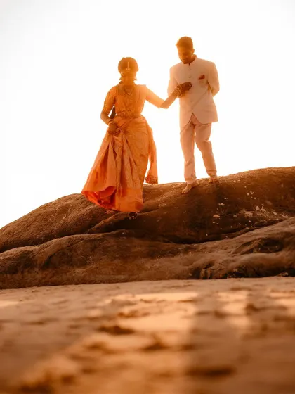 A couple walks hand-in-hand on a rocky shore during the golden hour. The warm sunset light creates a dreamy, romantic atmosphere perfect for a beachside wedding shoot.