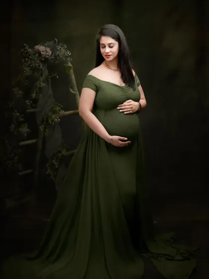 A full-length portrait in an olive green gown, set against a dark, moody backdrop with subtle floral elements. The lighting is carefully shaped to create a classic, painterly effect.
