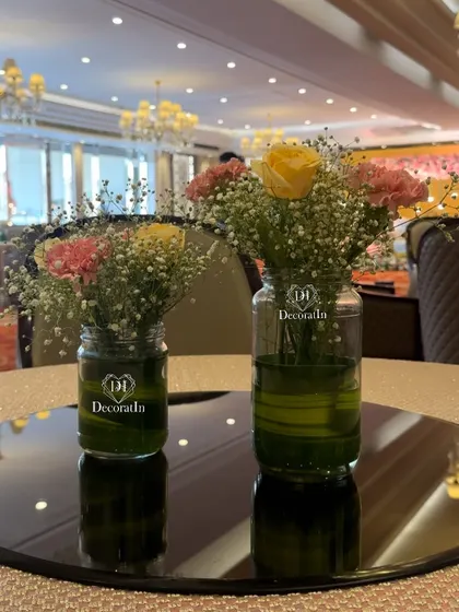 Small floral arrangements in glass jars on a mirrored base, creating a simple and reflective centerpiece for the guest tables.