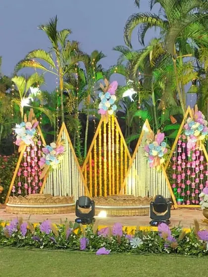 A grand outdoor Haldi stage setup. The triangular bamboo structures are decorated with floral arrangements and hanging strings, creating a beautiful backdrop against the natural greenery.