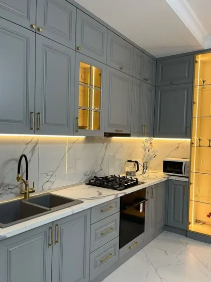 A beautiful grey kitchen in a classic style, featuring moulded cabinet doors and a tall, illuminated glass display case for showcasing fine glassware. The marble backsplash adds a touch of luxury.