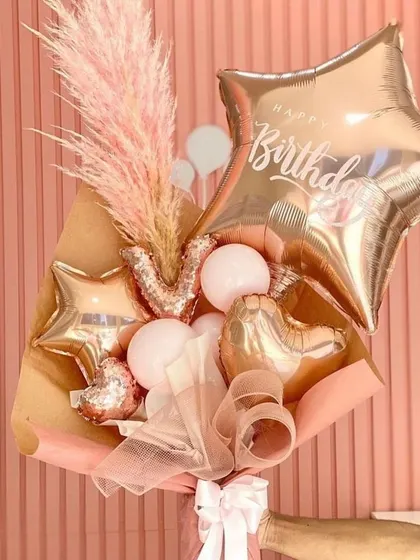 A glamorous birthday bouquet made with rose gold foil balloons, including stars, hearts, and a sequined letter 'M'. The addition of pink pampas grass adds a soft, feathery texture.