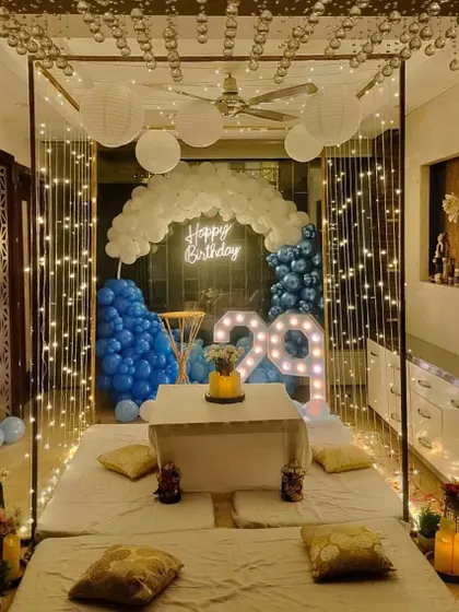 An intimate and cozy indoor birthday dinner setup. This features a cabana frame draped with fairy lights, low seating with plush cushions, and a balloon backdrop in shades of blue and white, perfect for a surprise celebration at home.