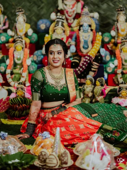 A vibrant portrait of the bride in a green and orange half-saree, seated amidst traditional dolls (Golu).